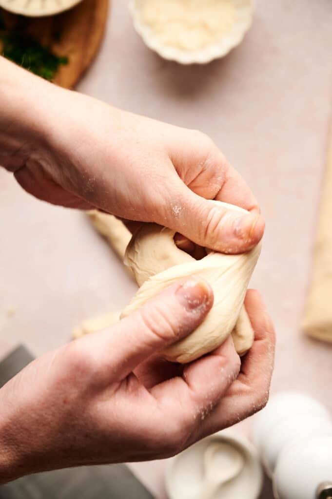 Close-up of hands kneading dough on a beige surface, perfecting the art of crafting garlic knots, with small dishes and ingredients blurred in the background.