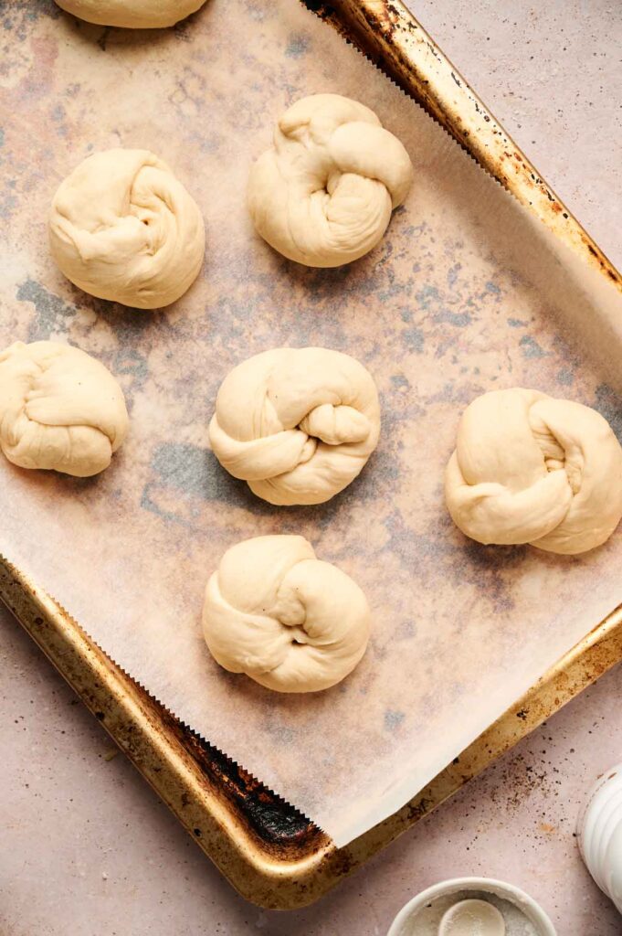 Six pieces of knotted garlic dough rest on a parchment-lined baking sheet, ready for baking.