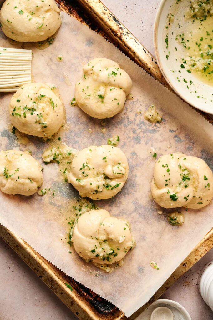 Garlic knots rest on a baking tray lined with parchment paper, glistening under a garlic and herb mixture. A nearby bowl holds the remaining savory blend alongside a pastry brush, ready for more delicious application.