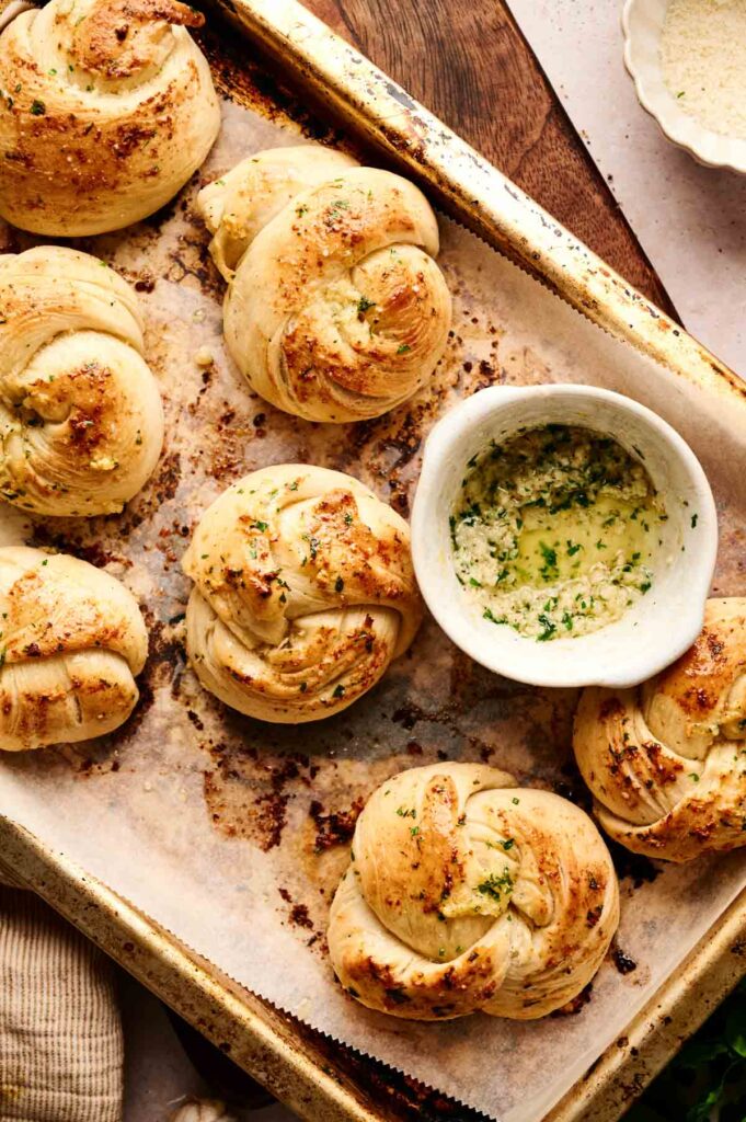Aromatic garlic knots rest on a parchment-lined tray, accompanied by a small bowl of velvety garlic butter.