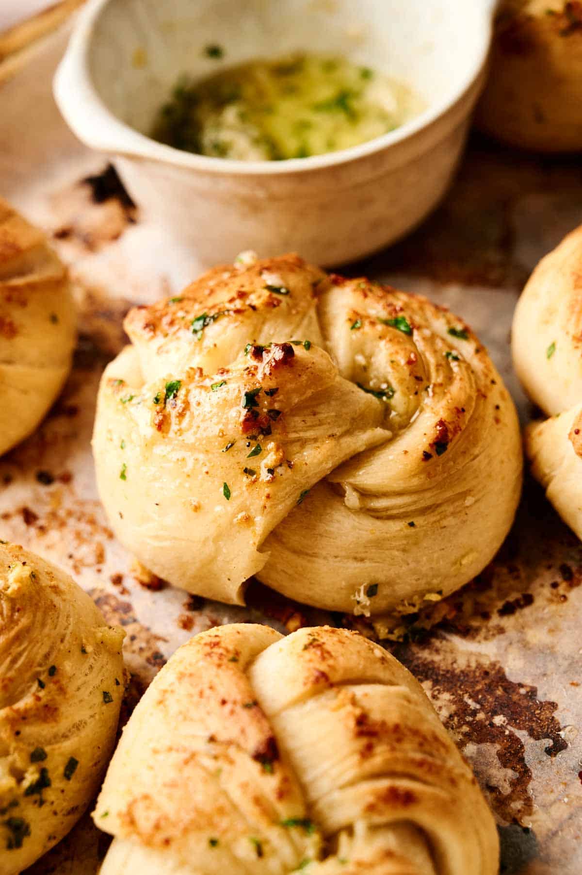 Freshly baked garlic knots rest on a baking sheet, enticingly paired with a small bowl of herb-infused oil in the background, ready to elevate your snacking experience.
