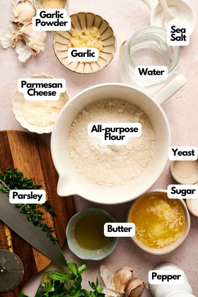 Ingredients for making garlic knots are arranged on a surface, including flour, garlic, parsley, butter, yeast, parmesan cheese, garlic powder, sugar, sea salt, pepper, and water.