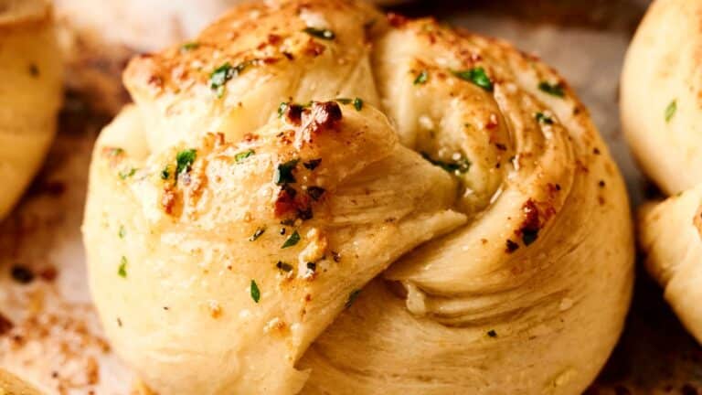A close-up of a baked garlic knot topped with chopped herbs and spices, displaying a golden-brown crust.