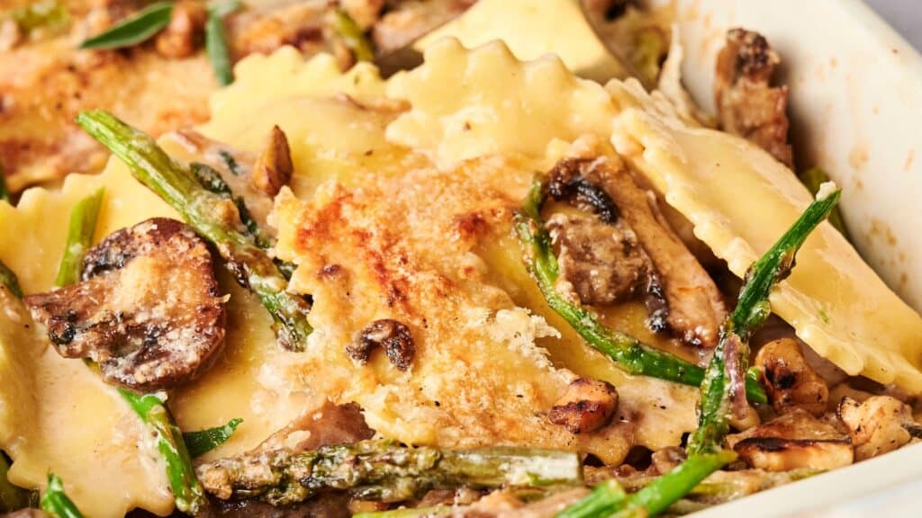 Close-up of baked ravioli with mushrooms and asparagus in a creamy sauce, topped with melted cheese.