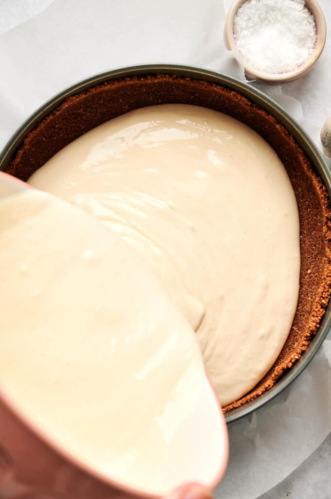 A creamy mixture is being poured into a round crust in a baking pan, with a small bowl of powdered sugar nearby.