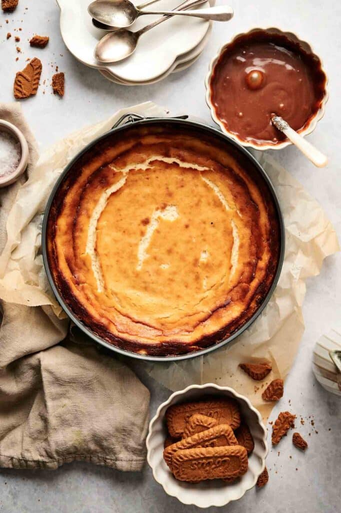 A baked cheesecake in a pan surrounded by empty plates, spoons, caramel sauce in a bowl, and Biscoff cookies on a light surface.