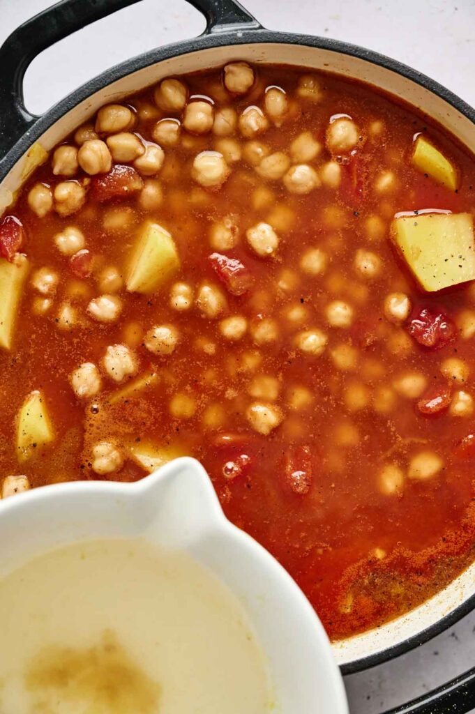 A pot of savory chickpea curry with potatoes and tomatoes in a rich red broth. In the foreground, a white bowl holds a light beige liquid, complementing the hearty dish.
