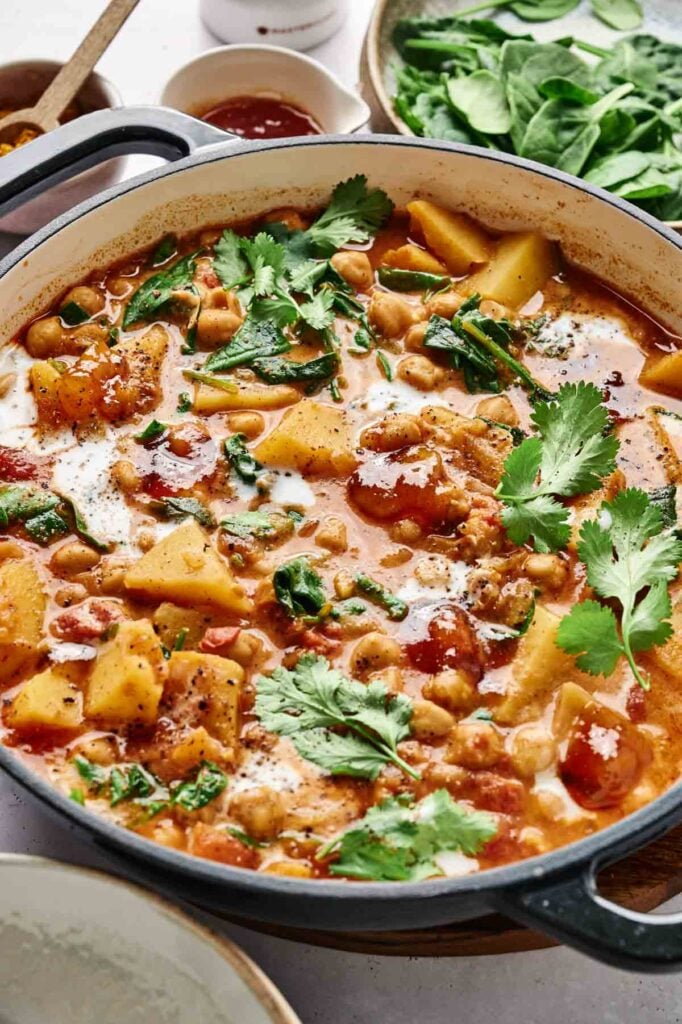 A pot of vibrant vegetable stew features chickpeas, potatoes, and tomatoes simmered in a delightful harmony reminiscent of chickpea curry. Garnished with fresh herbs, it’s surrounded by bowls of spinach leaves and aromatic spices for an enticing finish.