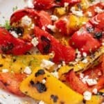 Grilled red and yellow bell peppers, perfectly charred, are topped with crumbled feta cheese on a white plate.