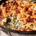 Cheesy spinach and artichoke dip in a black dish, with a portion missing, showing the creamy interior.