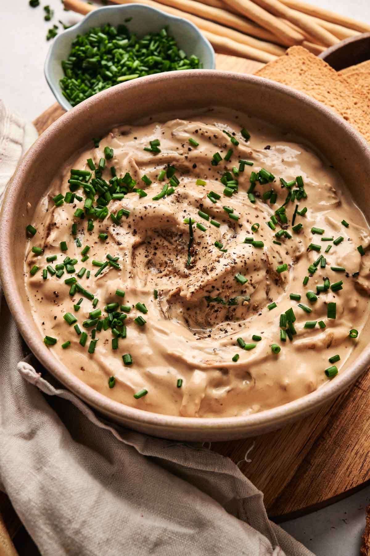 A bowl of creamy French onion dip garnished with chopped chives and black pepper, surrounded by crispy breadsticks.
