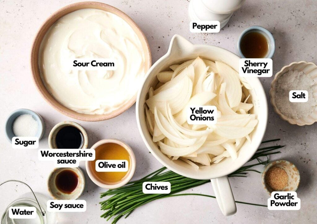 Overhead view of ingredients for French onion dip, including bowls of sour cream, yellow onions, pepper, sherry vinegar, salt, garlic powder, chives, olive oil, Worcestershire sauce, soy sauce, and water.
