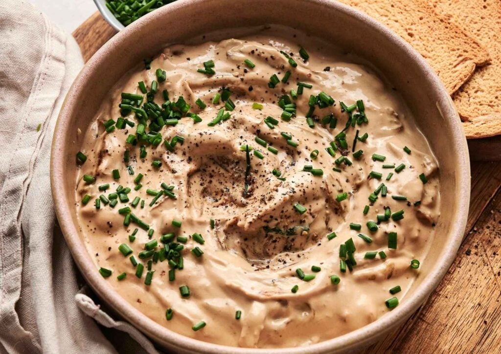 A bowl of creamy French onion dip garnished with chopped chives and black pepper, served with toasted bread.