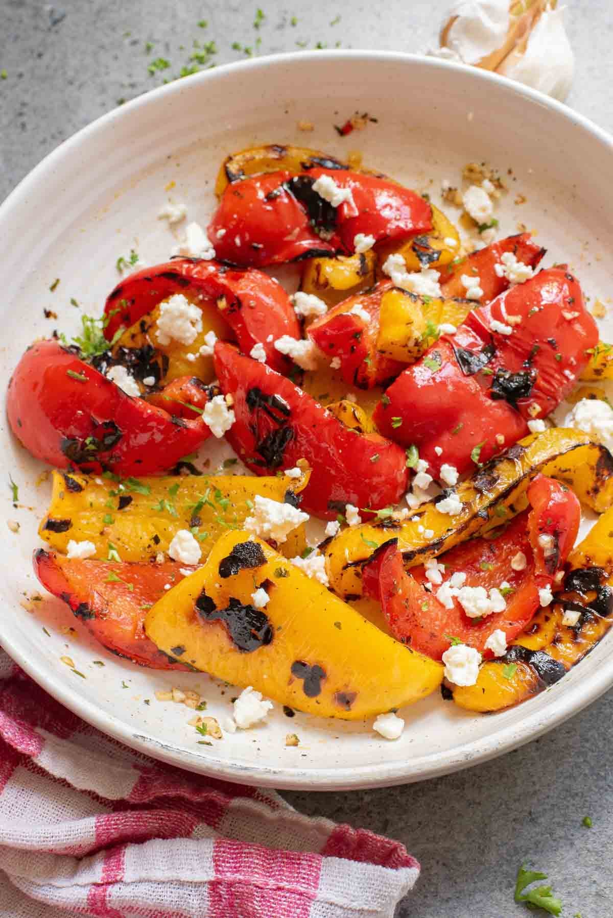 Grilled red and yellow bell peppers with crumbled feta and herbs rest on a white plate, garnished beautifully with parsley.