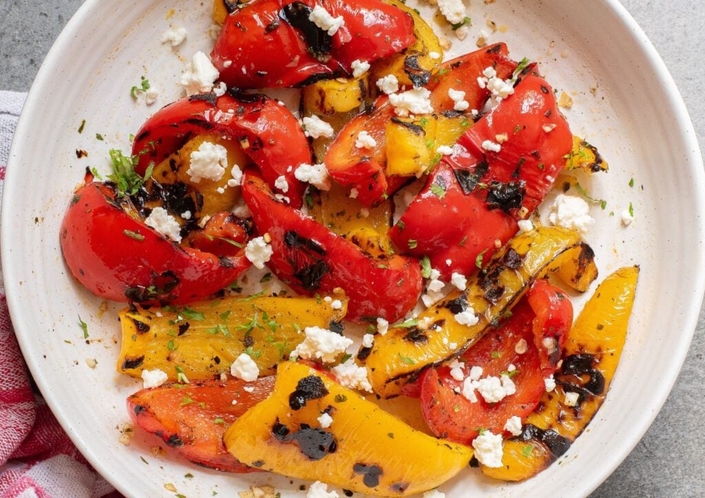 Grilled red and yellow bell pepper slices, bursting with flavor, are topped with crumbled feta cheese and herbs on a white plate—perfect for showcasing the smoky taste of grilled peppers.