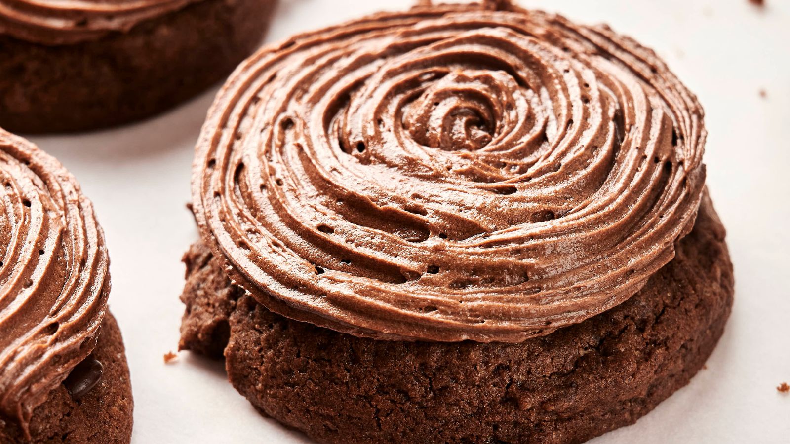 A close-up of a chocolate frosted cookie with swirl icing on a white surface, reminiscent of Copycat Crumbl Chocolate Cake Cookies and surrounded by crumbs.