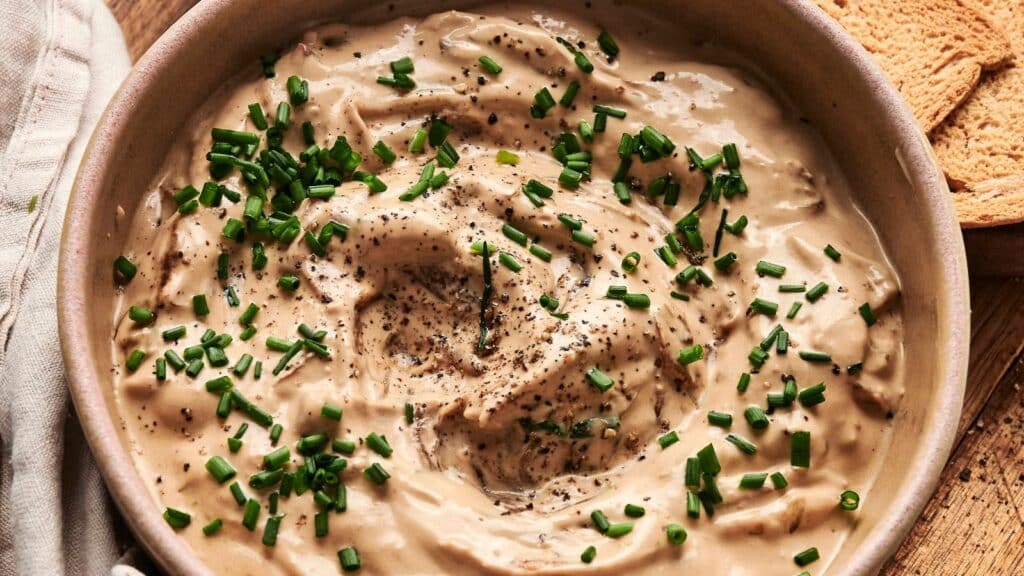 Bowl of creamy dip topped with chopped chives and black pepper, next to pieces of toasted bread.