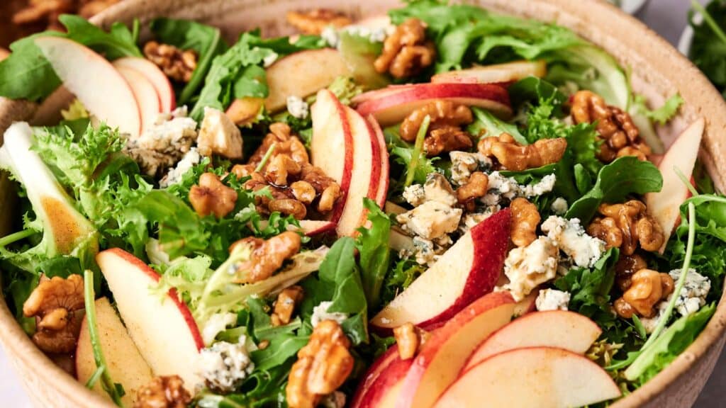 A wooden bowl filled with a mixed salad of fresh greens, apple slices, walnuts, and crumbled blue cheese.