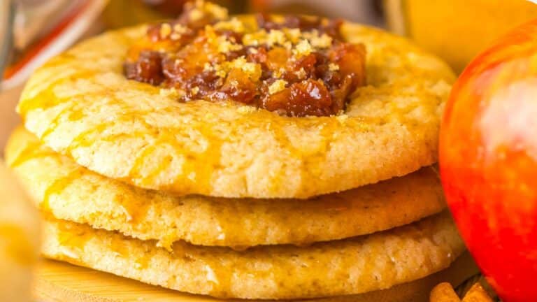 Close-up of three stacked cookies topped with a chunky apple filling, surrounded by a whole apple and an orange in the background.