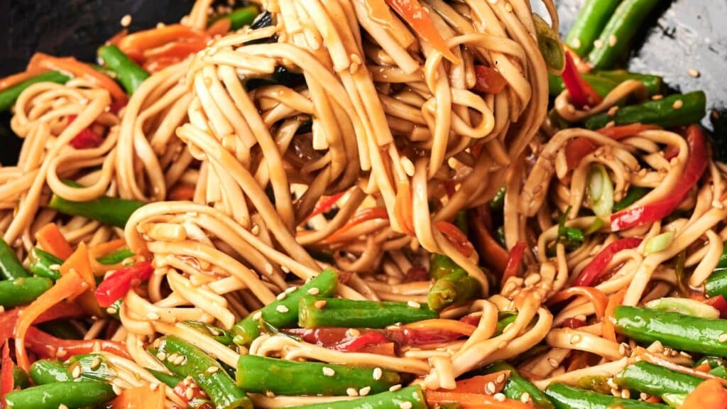 Stir-fried noodles with green beans, red bell peppers, and carrots, garnished with sesame seeds.