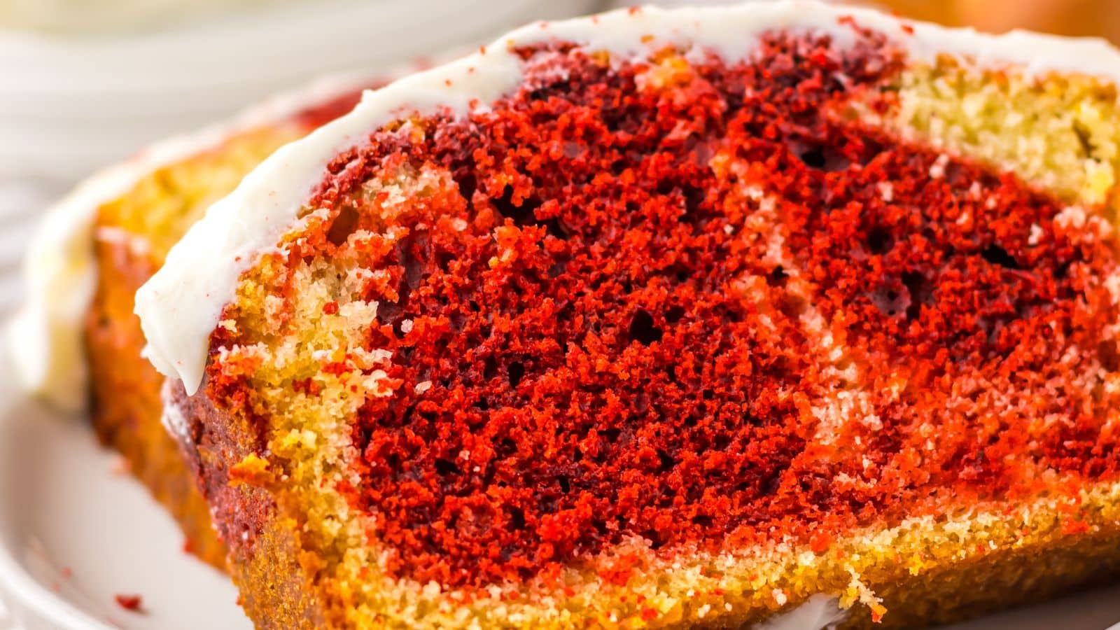 A slice of red and yellow marbled cake, reminiscent of a copycat Starbucks Red Velvet Loaf, is elegantly topped with white icing on a plate.