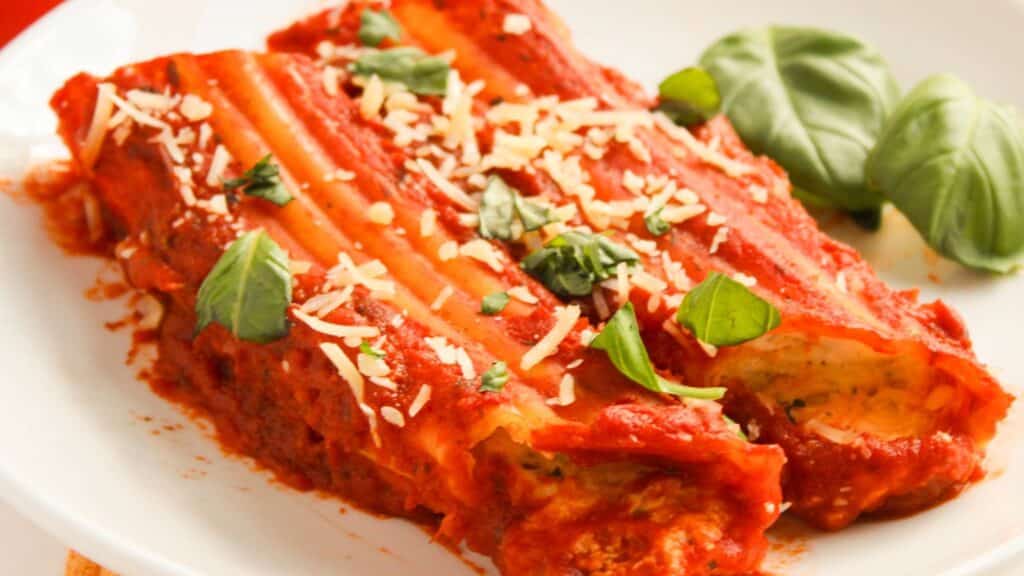Plate of cannelloni topped with red tomato sauce, shredded cheese, and fresh basil leaves.