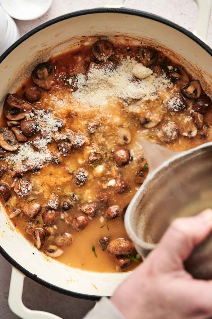 A person pours liquid into a pot containing ingredients for a delectable mushroom pasta, including mushrooms, tomato sauce, and grated cheese.