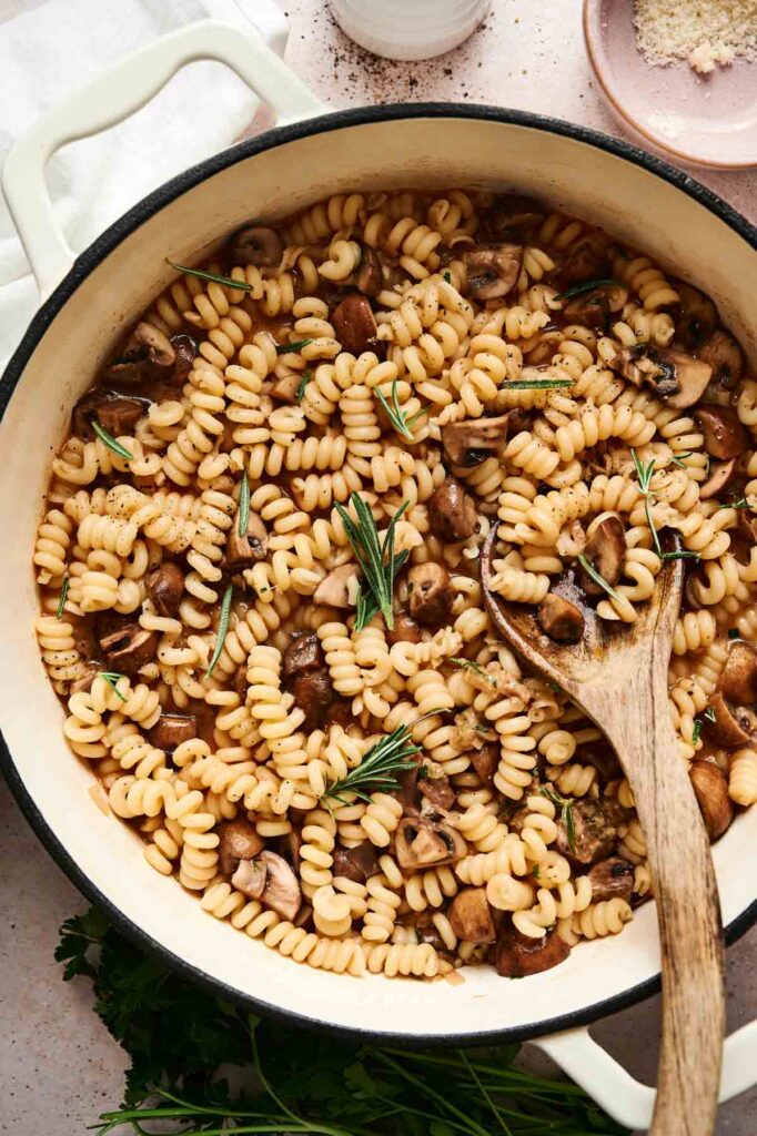 A pot of delightful mushroom pasta with spirals and rosemary, stirred gently with a wooden spoon on a light surface.