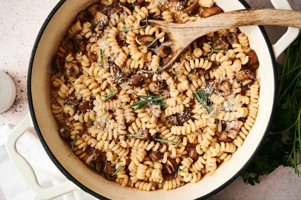 A pot of mushroom pasta with spirals, adorned with grated cheese and rosemary, has a wooden spoon nestled inside.