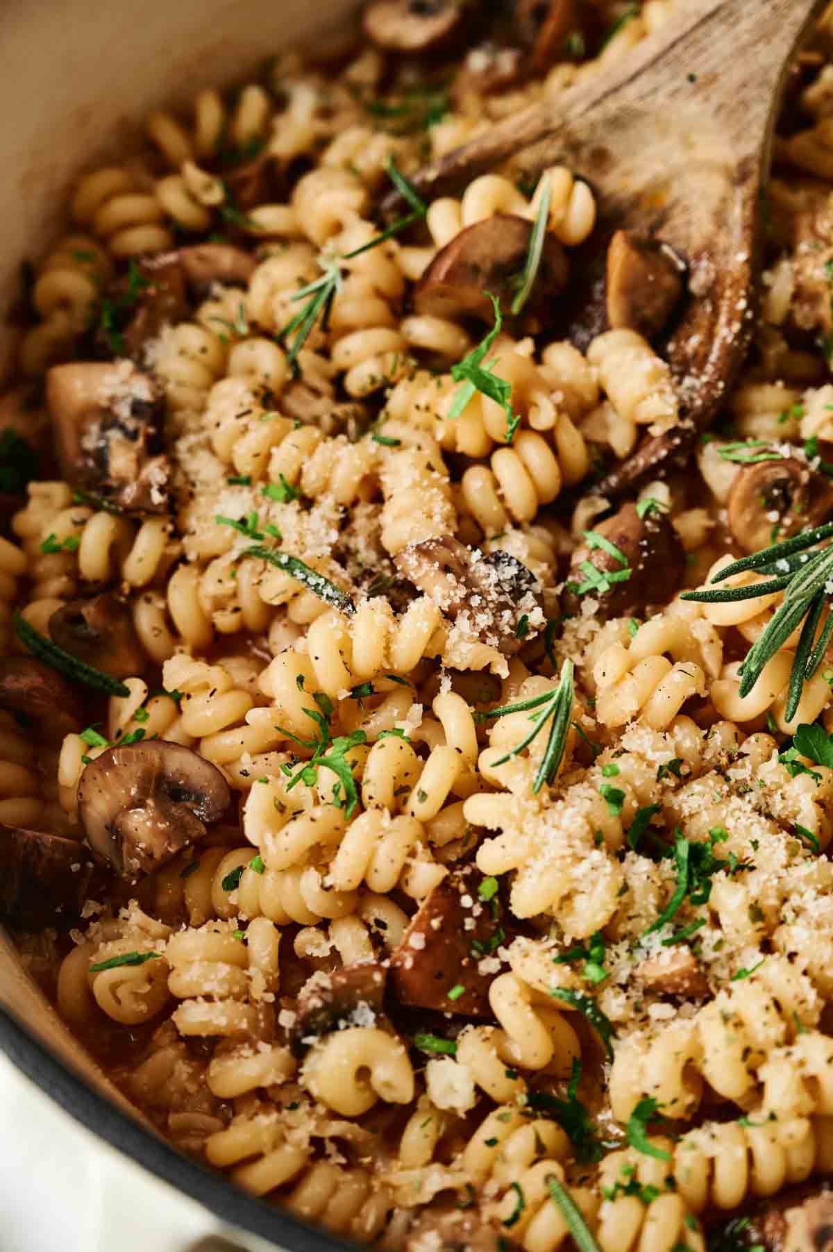 Close-up of mushroom pasta spirals mixed with mushrooms, garnished with grated cheese, fresh herbs, and a rosemary sprig, in a pan with a wooden spoon.