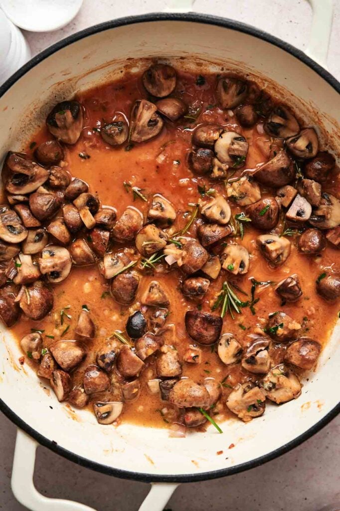 A pot of sliced mushrooms in a rich tomato sauce, creating a delectable base for mushroom pasta, is elegantly garnished with rosemary sprigs.