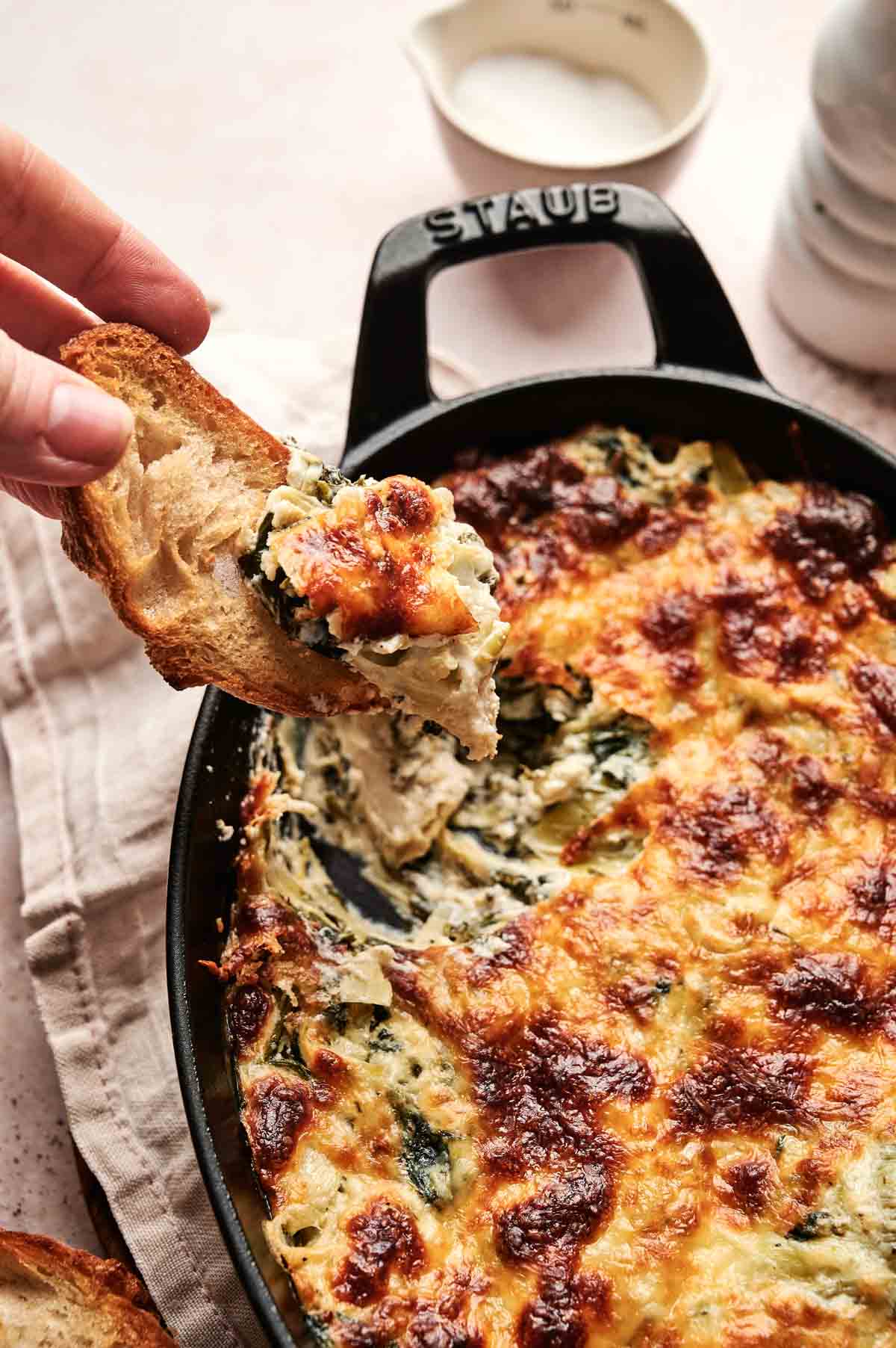 A hand holding a slice of bread topped with creamy spinach and cheese dip, above a black Staub dish containing more of the baked dip.