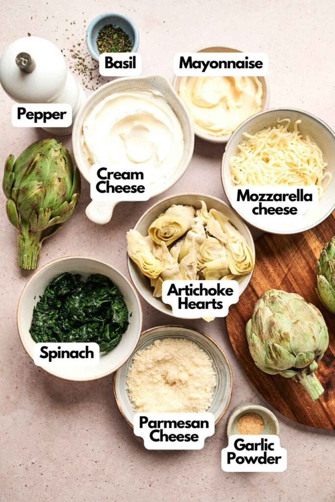 Ingredients for a dish are arranged on a table, including artichokes, spices, cream cheese, mayonnaise, mozzarella cheese, spinach, Parmesan cheese, and garlic powder.