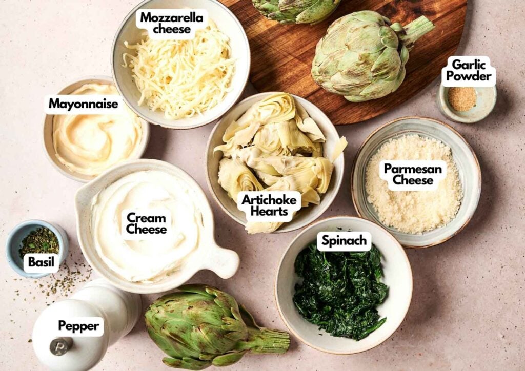 Bowls containing mozzarella cheese, garlic powder, mayonnaise, cream cheese, artichoke hearts, parmesan cheese, spinach, basil, and pepper are arranged on a countertop with fresh artichokes—perfect ingredients for a delicious spinach and artichoke dip.