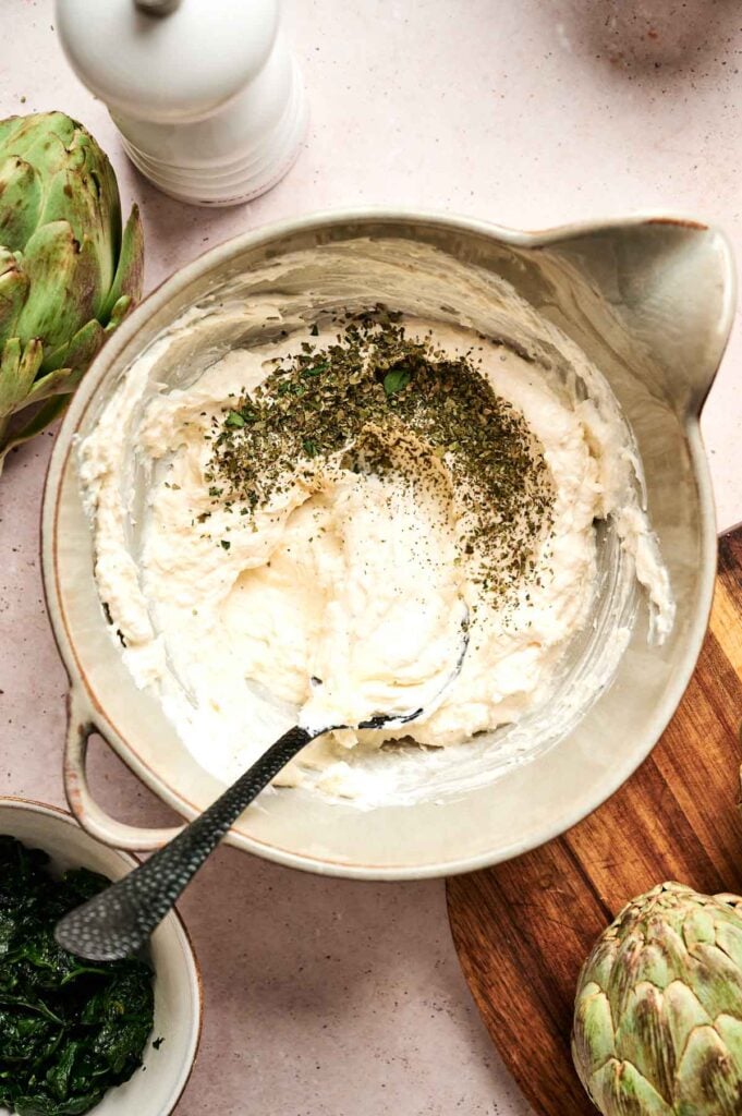 Bowl of creamy mixture with herbs, surrounded by artichokes and a bowl of greens on a light-colored surface.