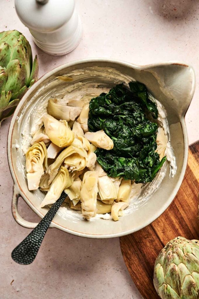 A bowl containing spinach and artichoke hearts mixed with creamy white sauce, with a spoon inside. Artichokes are nearby on a wooden surface.