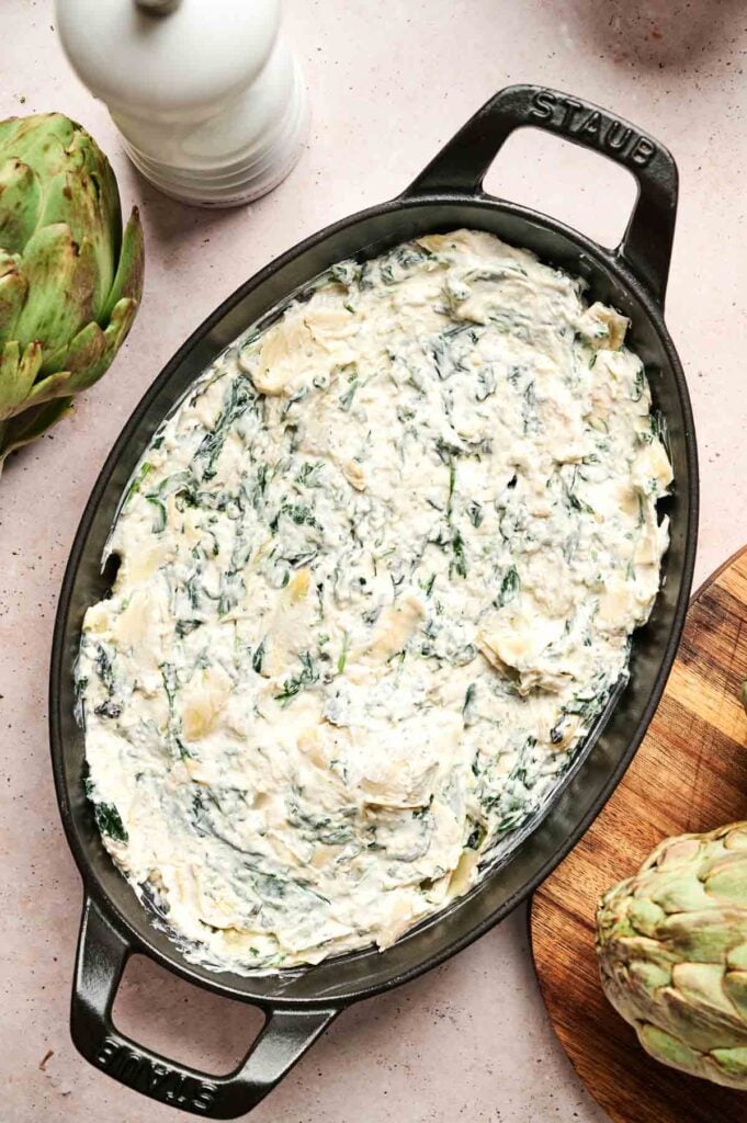Oval dish with creamy spinach artichoke dip, surrounded by artichokes and a pepper mill.
