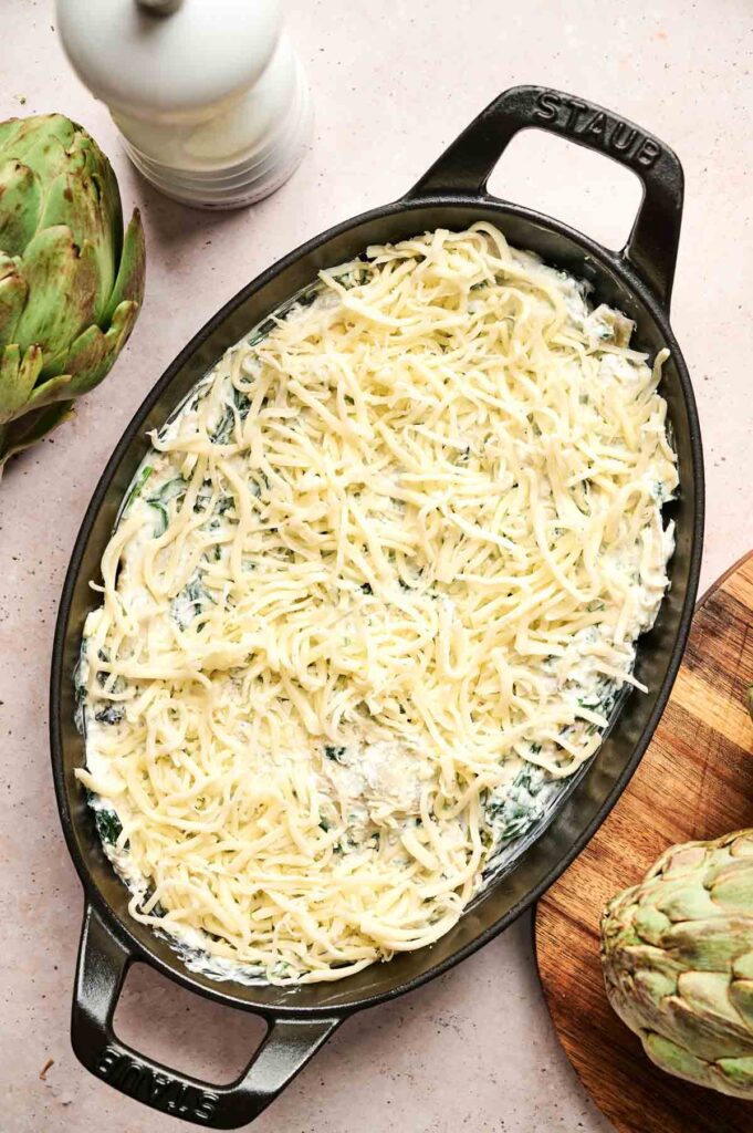 Oval baking dish filled with uncooked creamy spinach artichoke dip topped with shredded cheese, placed on a light surface with a whole artichoke and a pepper grinder nearby.