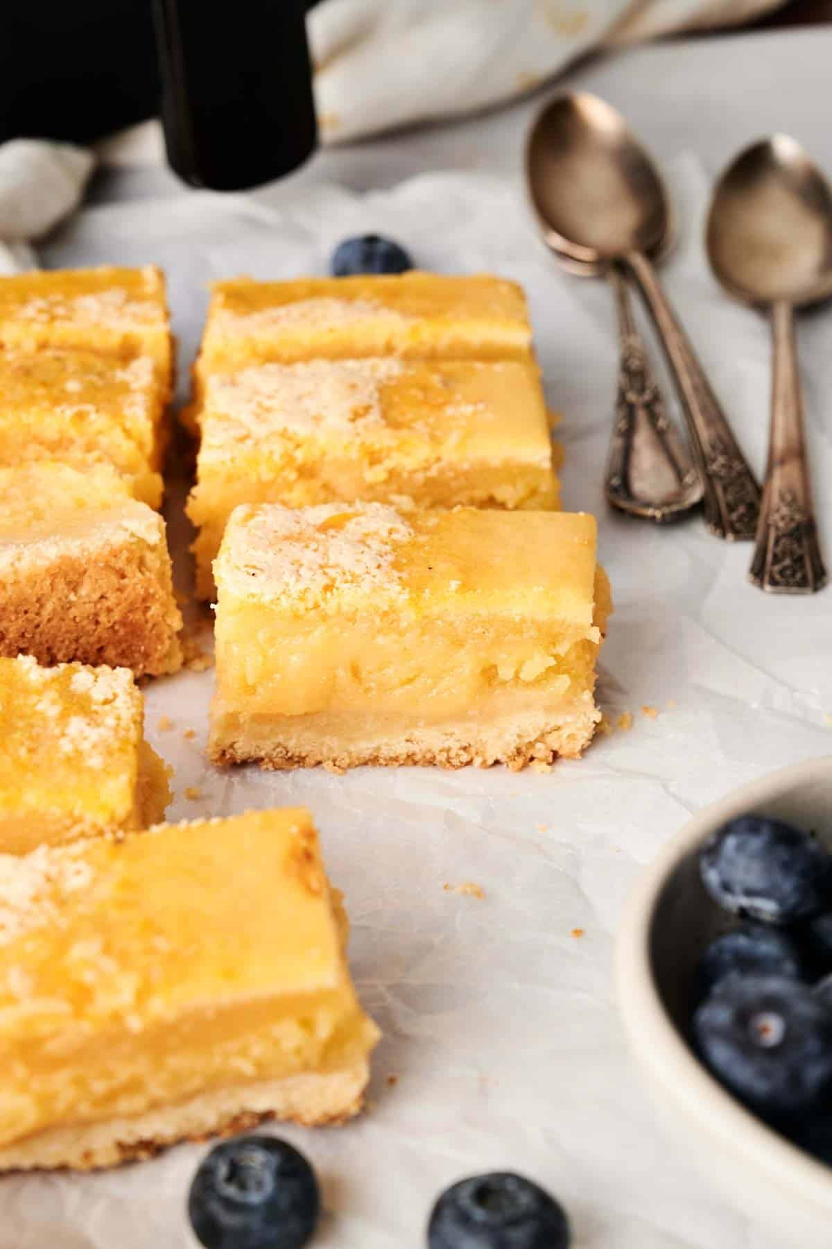 Air Fryer Lemon Bars rest on a parchment-lined surface, accompanied by three antique spoons and a bowl of blueberries nearby.