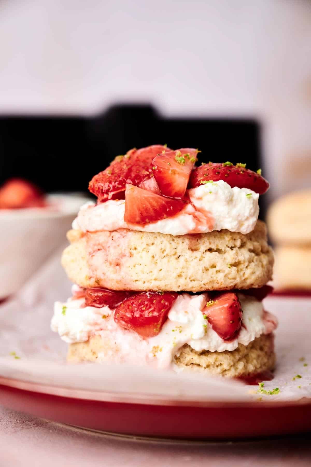 An air fryer strawberry shortcake with two biscuit layers, topped with whipped cream and sliced strawberries, served on a red plate.