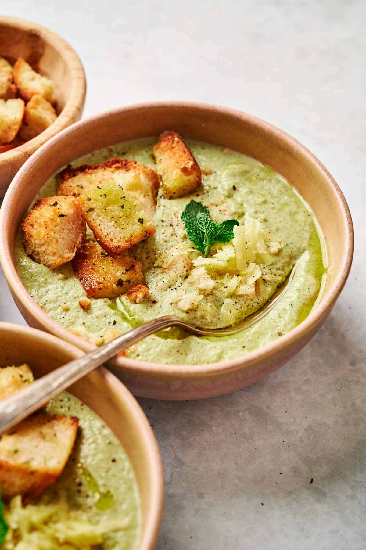 A bowl of creamy Air Fryer Broccoli Soup, crowned with croutons, grated cheese, and a mint sprig, a spoon resting within. Another bowl waiting in the background showcases its own stash of crispy croutons.