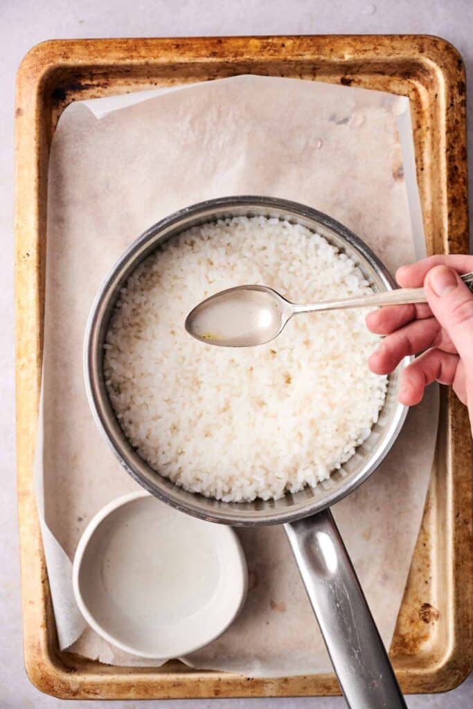 A person holds a spoon over a pot of crispy rice, placed on a baking tray with parchment paper, next to an empty bowl.
