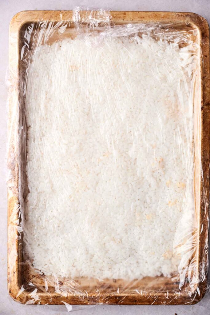 Cooked rice spread on a baking sheet, waiting to transform into crispy rice, covered with plastic wrap.