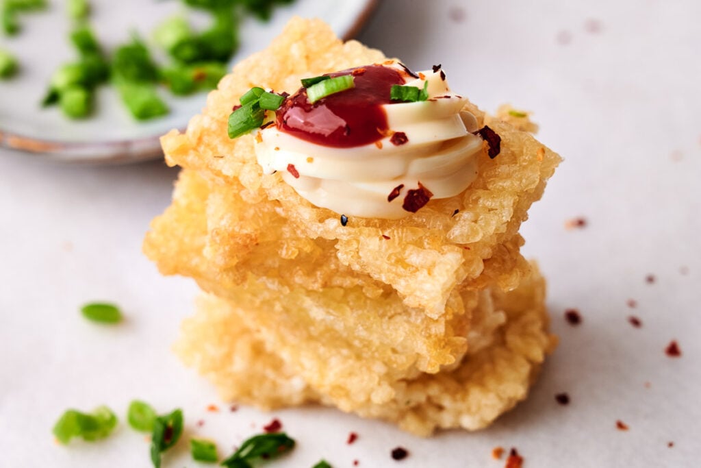 Three stacked crispy rice cakes, featuring the irresistible crunch of crispy rice, are topped with mayonnaise, red sauce, chopped green onions, and a sprinkle of red pepper flakes on a white surface.