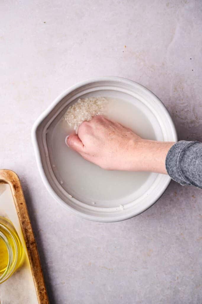 A hand gently rinses rice in a white bowl filled with water on the countertop, preparing the grains for a deliciously crispy rice dish. A wooden board with a jar of oil is visible on the left, ready to add flavor and texture.