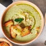 A bowl of vibrant Air Fryer Broccoli Soup, garnished with croutons, grated cheese, a mint leaf, and a drizzle of olive oil.