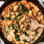 A skillet filled with creamy "Marry Me" butter bean stew featuring spinach, sun-dried tomatoes, and a wooden spoon.