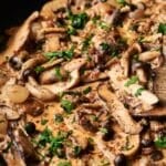 Close-up of a creamy mushroom stroganoff sauce with slices of various mushrooms, chopped parsley, and black pepper, elegantly stirred with a wooden spoon.