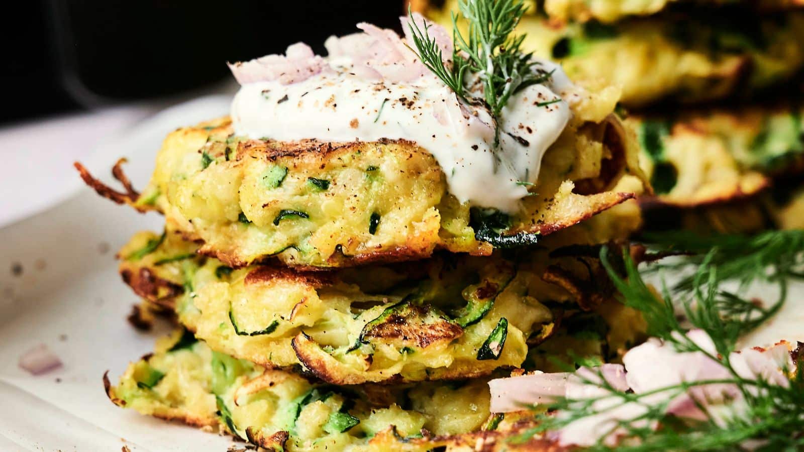 A stack of zucchini fritters topped with sour cream, dill, and chopped onions on a plate.