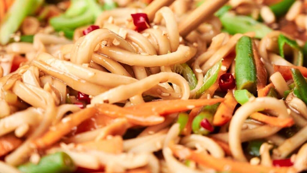 Close-up of stir-fried noodles with vegetables, including carrots, green beans, and chopped red peppers, garnished with sesame seeds. Chopsticks are picking up some noodles.